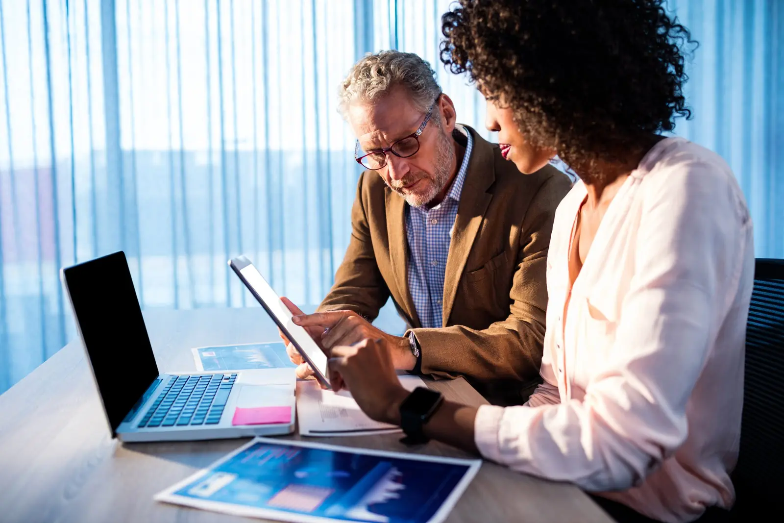 Two colleagues reviewing work together on a tablet beside an open laptop
